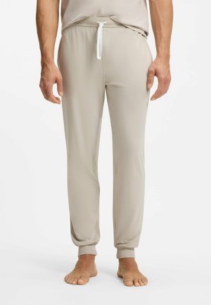 Man die beige joggingbroek met trekkoord en bijpassend shirt draagt, blootsvoets staand tegen een witte achtergrond.