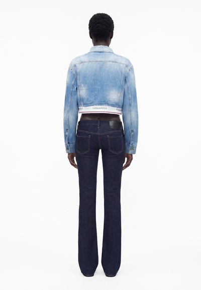 Άτομο από πίσω με cropped ανοιχτό μπλε denim μπουφάν και σκούρα μπλε παντελόνια καμπάνα σε απλό λευκό φόντο.