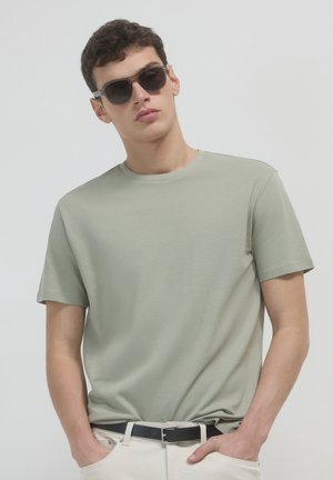 T-shirt vert clair texturé avec un col rond, associé à une ceinture noire et un pantalon crème. Le modèle porte des lunettes de soleil noires.