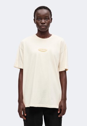 Ung person med kort hår iført en lysebeige Patagonia T-shirt og sorte bukser, stående mod en ensfarvet lys baggrund.