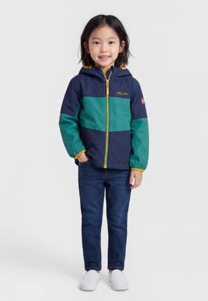Bambino con capelli neri che indossa una giacca con cappuccio blu navy e verde, jeans blu e scarpe slip-on bianche, in piedi con le mani in tasca.