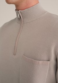 Hellgrauer Pullover mit einem hohen Rippkragen und einem Viertelreißverschluss, ausgestattet mit einer aufgesetzten Tasche auf der linken Brust aus passendem Stoff.