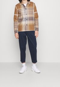 Camisa de botones en un patrón de cuadros marrón y beige, con mangas largas y cuello. Combinada con pantalones azul marino y zapatillas blancas.