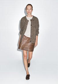 Braun strukturierte Strickjacke, beiges Oberteil, brauner Leder-Minirock und dunkelbraune Schuhe. Model läuft vor schlichtem Hintergrund.