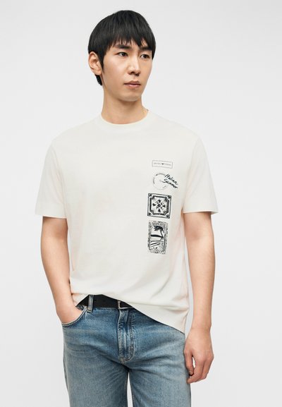 Νεαρός άνδρας που φοράει λευκό μπλουζάκι με μαύρα σχέδια και μπλε τζιν, στέκεται με το ένα χέρι στην τσέπη σε απλό φόντο.