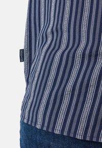 Marineblauwe shirt met witte verticale strepen, met een gedetailleerd patroon en een label met de tekst "SEASIDE" aan de onderrand. Getextureerde stof.