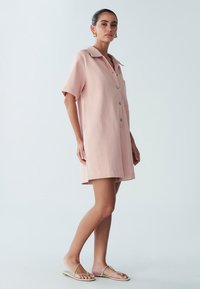 Svetlo roza obleka s kratkimi rokavi, z gumbi spredaj in ovratnikom, z enim žepom, teksturirano tkanino ter stranskimi razporki za lažje gibanje.