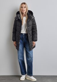 Svart puffjacka med huva och quiltat mönster, med tryckknappar och dragkedja. Matchad med blå löst sittande jeans och vita sneakers.