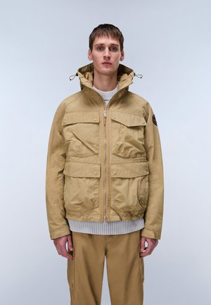 Mand står med fronten mod kamera iført en beige hættejakke med fire frontlommer og matchende beige bukser.