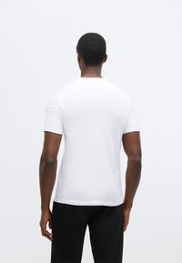 Biele tričko s krátkym rukávom z hladkej látky, s okrúhlym výstrihom a priliehavým strihom, zobrazené zozadu. Nižšie sú viditeľné čierne nohavice.