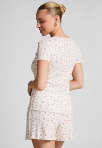 Korte pyjama set met bloemenpatroon in witte ribbed stof, met golvende randen aan mouwen en shorts. Roze bloemen contrasteren met de achtergrond.