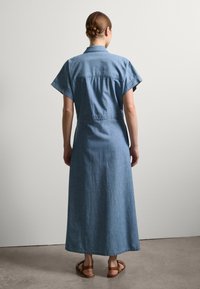 Abito lungo in denim di colore azzurro chiaro con maniche corte, colletto e vita cinchata. Presenta una vestibilità ampia con gonna dritta e sandali marroni.