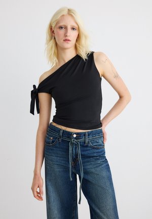 Blond žena v čiernom topu na jedno rameno s mašľou a vysokými modrými džínsami so zviazaným opaskom, stojaca pred jednoduchým pozadím.