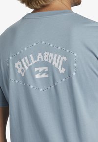 Svetle modré bavlněné tričko s velkým bílým logem "BILLABONG" na zadní straně, orámované květinovými akcenty ve tvaru hvězdy. Krátké rukávy, kulatý výstřih.