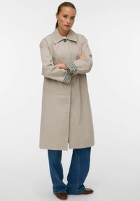 Beige trench coat kraega ja ruuduliste varrukatega, nupupuclosinguga, kantud laia jala siniste teksade ja pruunide kingadega.