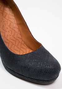 Zapato de tacón azul marino con un patrón texturizado y punta redonda. El interior es de cuero color marrón claro con un diseño geométrico.