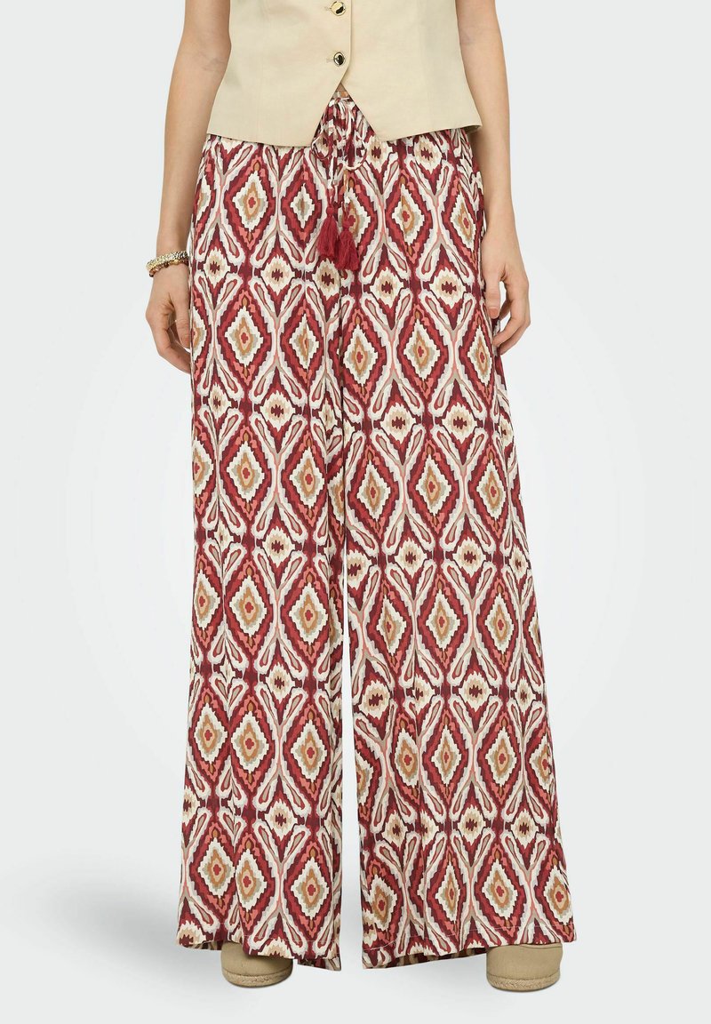 Femme portant un pantalon à jambes larges avec un motif géométrique de diamants rouge, blanc et beige et un haut sans manches beige.
