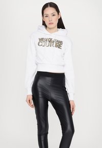 Hoodie bianco cropped con testo dorato "VERSACE JEANS COUTURE"; abbinato a leggings neri lucidi a vita alta. Finitura in tessuto liscia in tutto.