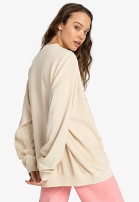 Beige oversized sweatshirt with long sleeves and ribbed cuffs, featuring a graphic design on the back. Worn with pink pants.