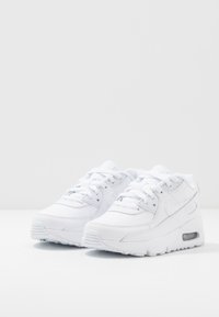 Zapatillas blancas con parte superior de cuero sintético, punta redonda, sistema de cierre con cordones, cuello acolchado y amortiguación de aire visible en el talón.