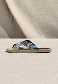 Επίπεδη σάνδαλα με πλεκτή σόλα και μπλε υφασμάτινες λουρίδες με ζικ-ζακ που διασταυρώνονται πάνω στο πόδι, παρουσιασμένη σε φωτεινή επιφάνεια με μπεζ φόντο.