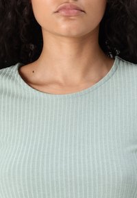 Närbild av en persons underansikte, nacke och övre bröst, iförd en ribbad ljusgrön topp med lockigt mörkt hår synligt.