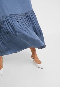 Blå plisserad kjol som flödar över benen som bär vita spetsiga slingback-klackar på ett vitt golv.