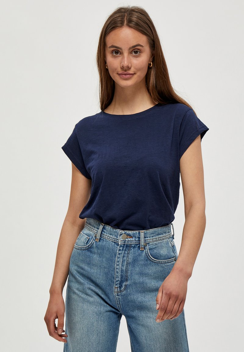Młoda kobieta z długimi brązowymi włosami, ubrana w granatowy t-shirt z krótkim rękawem i wysokim stanem niebieskie dżinsy, stoi na jednolitym tle.