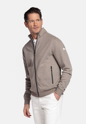 Man met kort haar, gekleed in een beige jas met rits en witte broek, kijkt naar links met één hand in zijn zak.