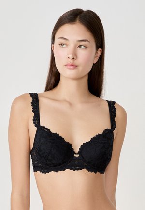 Reggiseno in pizzo nero con motivo floreale e bordi scallop. Presenta spalline regolabili e un scollo profondo con un ponte sotto le coppe.