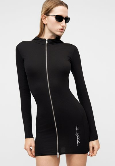 Γυναίκα με εφαρμοστό μαύρο μίνι φόρεμα με φερμουάρ και μακριά μανίκια, λευκό κείμενο κοντά στο τελείωμα και μαύρα γωνιώδη γυαλιά ηλίου, που στέκεται μπροστά σε λευκό φόντο.