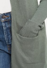 Cardigan verde din material tricotat moale, având un buzunar mare și mansete cu nervuri, asortat cu blugi albaștri.