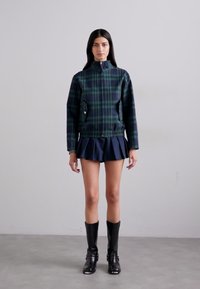 Giacca imbottita a quadri blu-verde con colletto alto, tasche frontali e orlo elasticizzato, abbinata a una gonna plissettata blu navy e stivali neri al ginocchio.