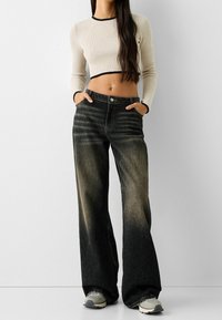 Svarta vida jeans med blekta detaljer, parat med en krämfärgad ribbad crop top med svarta kanter. Modellen står med händerna i fickorna.