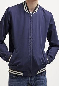 Veste aviateur bleu marine avec une fermeture éclair à l'avant, un col et des poignets rayés, et des poches latérales. Le matériau semble lisse et léger.