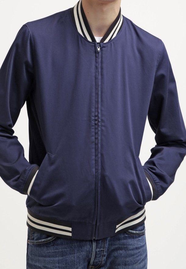 Veste aviateur bleu marine avec une fermeture éclair à l'avant, un col et des poignets rayés, et des poches latérales. Le matériau semble lisse et léger.