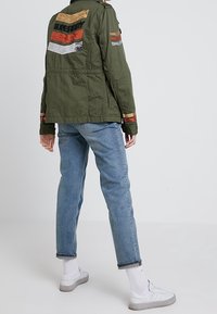 Chaqueta verde oliva bordada con rayas multicolores en la parte trasera y las mangas, combinada con vaqueros azul claro y zapatillas blancas.