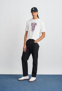 Hvid T-shirt med farverigt grafisk design, sorte jeans og hvide sneakers. Mørk baseballcap med logo. Glat stof og afslappet pasform.