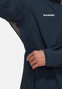 Veste technique bleu marine avec manches longues, fermeture à glissière et logo 'Mammut'. Elle possède un tissu texturé et des poignets ajustables.