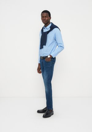 Homme portant une chemise bleu clair, un pull sombre drapé sur les épaules, un jean bleu foncé, des mocassins noirs, debout devant un fond blanc.