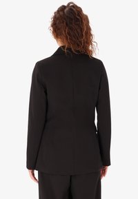 Blazer nero strutturato con colletto a punta, vestibilità aderente, tessuto liscio e maniche leggermente affusolate, vista posteriore e dettagli di cucitura.