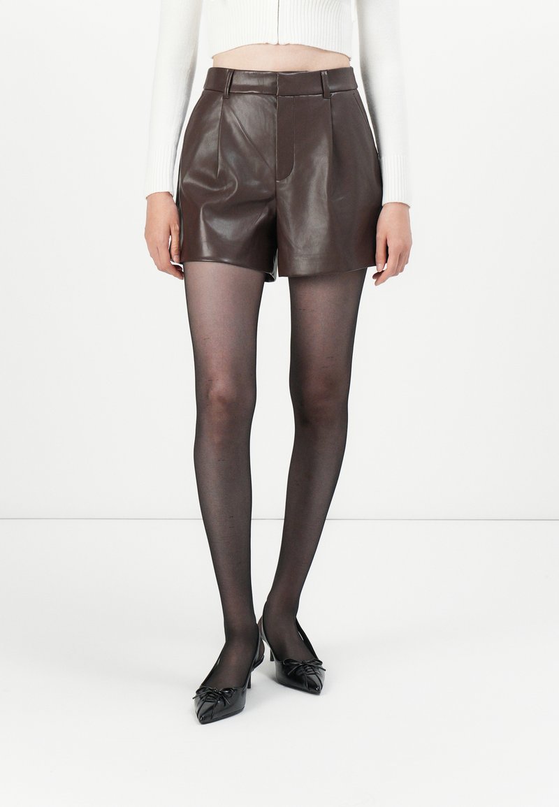 Brown leather high-waisted shorts with pleats, paired with sheer black tights and black pointed flats featuring decorative bows.