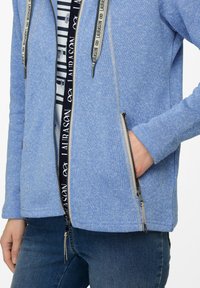 Blå, texturerad zip-up jacka med kontrasterande dragkedjedetaljer och justerbara snörningar. Har sidofickor och en huva.