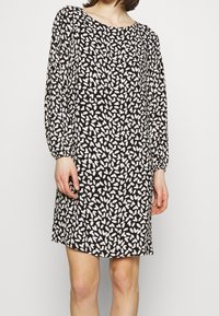 Nainen, jolla on päällään musta pitkähihaista mekko, jossa on valkoinen abstrakti terälehtikuvio, seisoo yksinkertaista vaaleaa taustaa vasten.