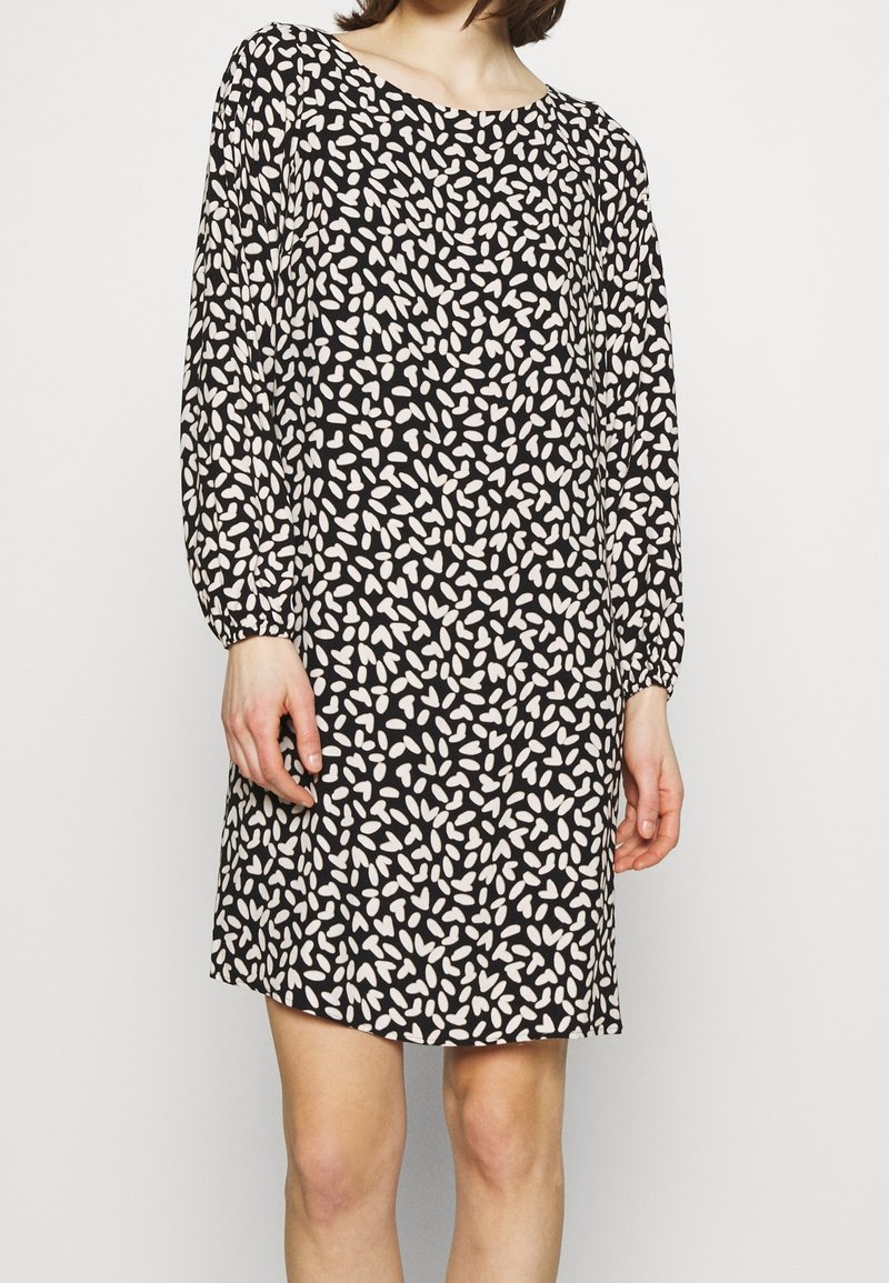 Nainen, jolla on päällään musta pitkähihaista mekko, jossa on valkoinen abstrakti terälehtikuvio, seisoo yksinkertaista vaaleaa taustaa vasten.