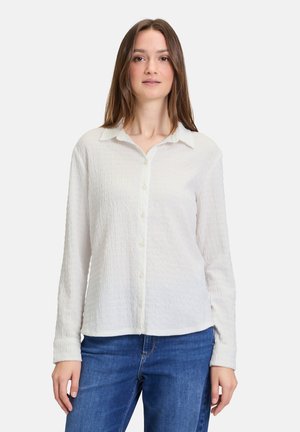 Weißes Hemd mit strukturierter Oberfläche, langen Ärmeln und spitzem Kragen, kombiniert mit blauen Jeans, präsentiert an einem Modell.