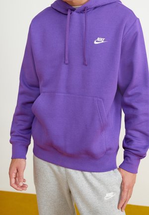 Osoba ubrana w fioletową bluzę z kapturem Nike z przednią kieszenią i szare spodnie dresowe Nike, stoi z rękami swobodnie opuszczonymi wzdłuż ciała.