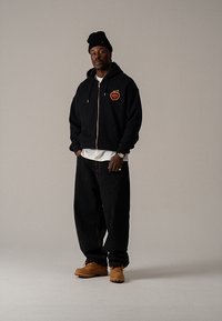 Felpa nera con zip e logo della mela, indossata sopra una maglietta bianca. Jeans larghi e scuri e stivali marroni. La modella indossa anche un berretto nero.