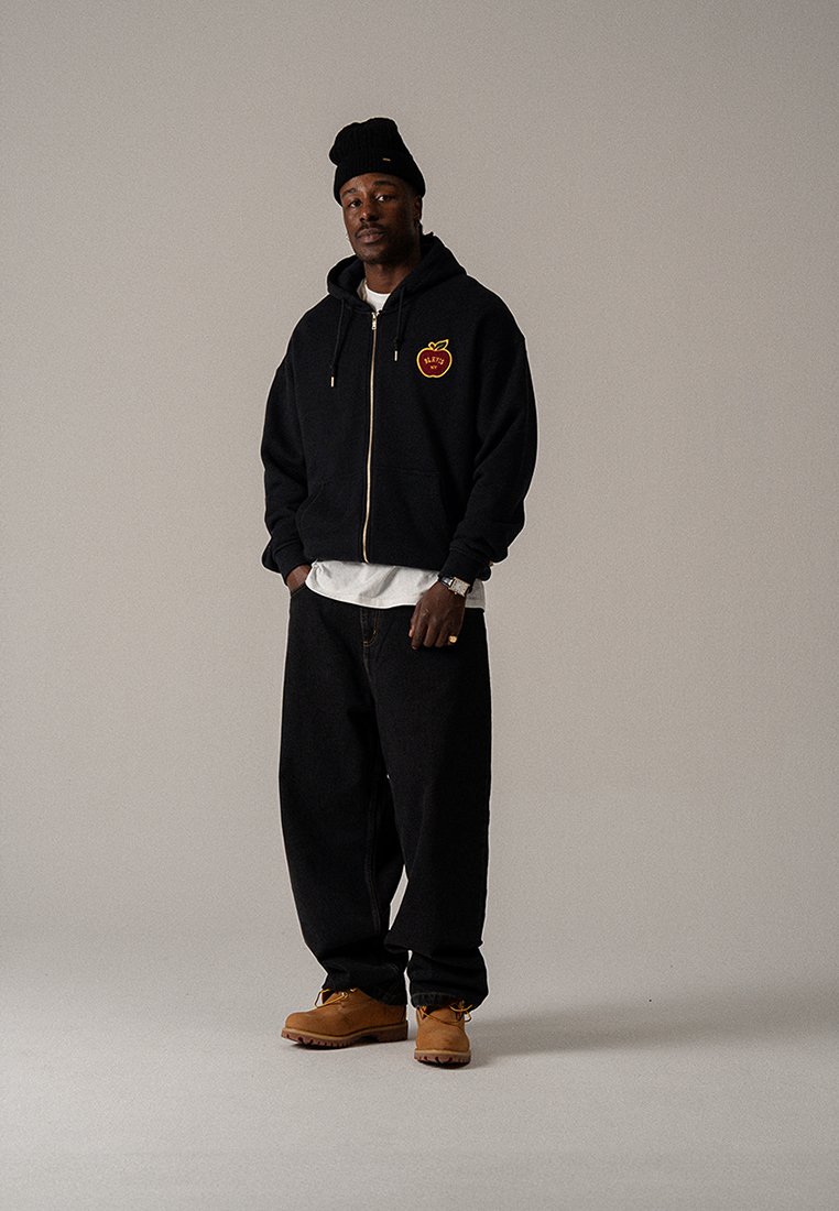 Felpa nera con zip e logo della mela, indossata sopra una maglietta bianca. Jeans larghi e scuri e stivali marroni. La modella indossa anche un berretto nero.