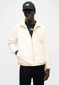 Άνδρας με εκρού φλις μπουφάν, μαύρο T-shirt, μαύρο καπέλο με λογότυπο The North Face, στέκεται με το ένα χέρι στην τσέπη του μπουφάν πάνω σε λευκό φόντο.